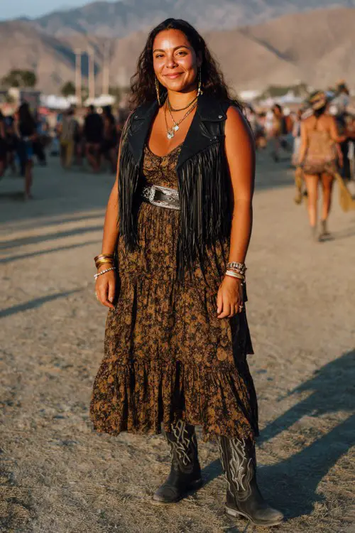 A curvy woman wears embroidered black cowboy boots with a tiered boho maxi dress and wide western belt, layered with a fringe vest