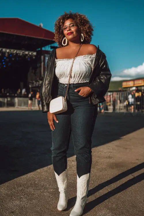 A plus-size woman wears white cowboy boots with tailored dark jeans, a fitted off-shoulder top and cropped leather jacket, finished with statement earrings and mini shoulder bag
