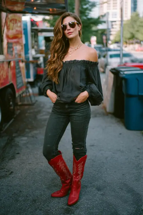 A woman over 30 wears dark skinny jeans styled with red cowboy boots, an off-shoulder blouse, and oversized sunglasses