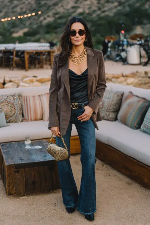 A woman over 30 wears sleek black cowboy boots with flared blue jeans, a silky camisole and tailored suede blazer, accessorized with layered gold chains and structured clutch