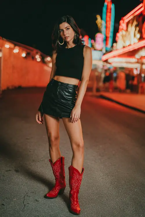 A woman wears bold red cowboy boots with a black leather mini skirt, fitted crop top, and oversized hoop earrings