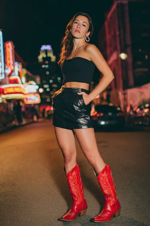 A woman wears bold red cowboy boots with a black leather mini skirt, fitted crop top, and oversized hoop earrings