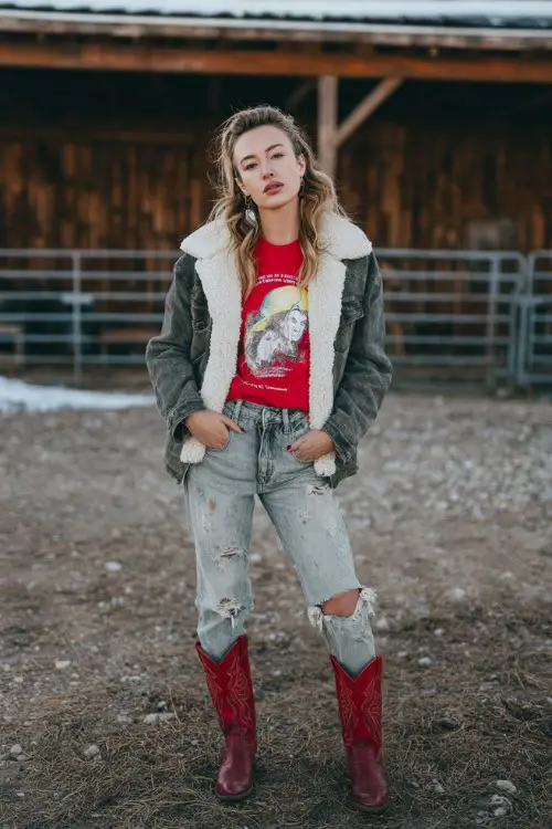 A woman wears distressed jeans with red cowboy boots, a shearling jacket, and a graphic tee layered underneath