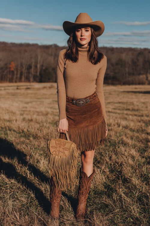 Woman in an open field with a concert stage behind her, wearing a fitted tan turtleneck sweater, brown suede fringe skirt, wide western belt, tan cowboy hat