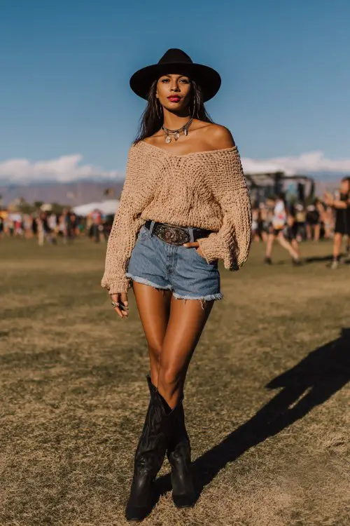Woman standing at an outdoor festival venue, wearing an oversized off-shoulder beige knit sweater, high-waisted denim cutoff shorts