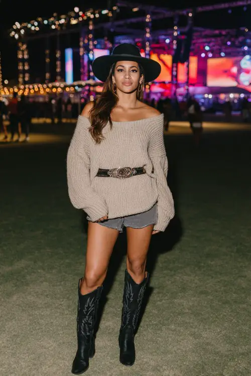Woman standing at an outdoor festival venue, wearing an oversized off-shoulder beige knit sweater, high-waisted denim cutoff shorts