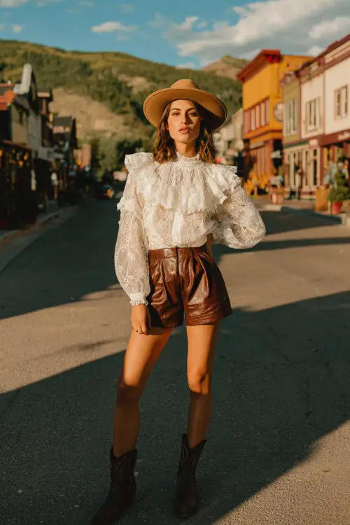 Woman wearing a white lace ruffled blouse, high-waisted brown leather shorts, tan wide-brim western hat, standing on a small western town street