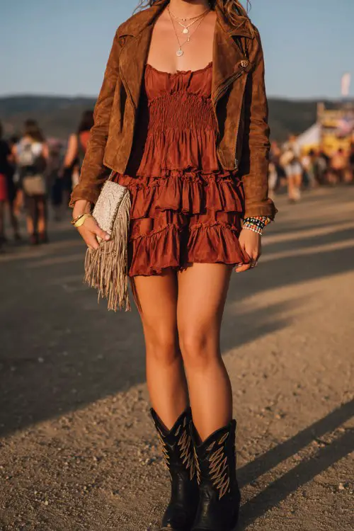 curvy woman wears black cowboy boots with a ruffled boho mini dress layered under a cropped suede jacket, accessorized with stacked bracelets and a fringe clutch