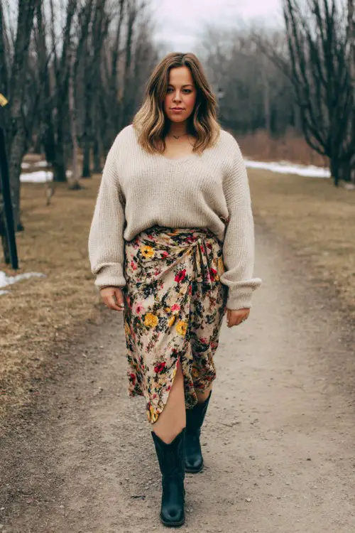 A plus size woman wears black cowboy boots with a soft beige oversized sweater and a floral wrap skirt
