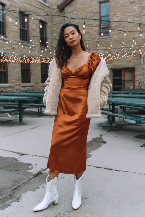 A woman over 30 wears a copper satin midi dress with puff sleeves and white cowboy boots, styled with a cropped faux fur jacket