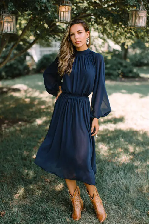 A woman over 30 wears a navy chiffon midi dress with long bishop sleeves and caramel cowboy boots, styled with pearl drop earrings