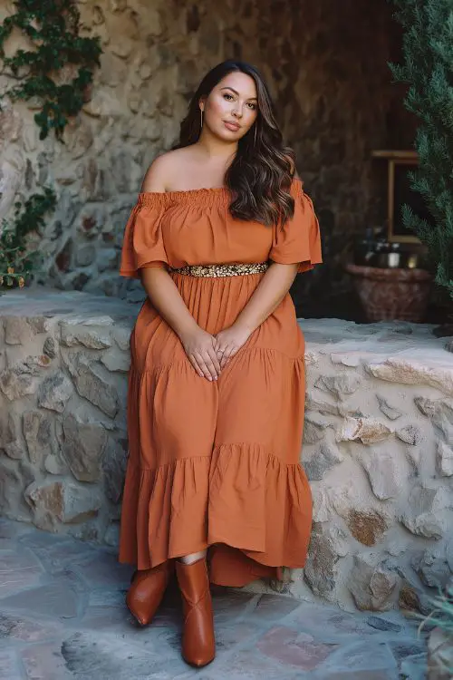 A curvy guest in a burnt orange off-the-shoulder maxi dress with a tiered skirt and belted waist, paired with heeled boots and soft waves in fall wedding