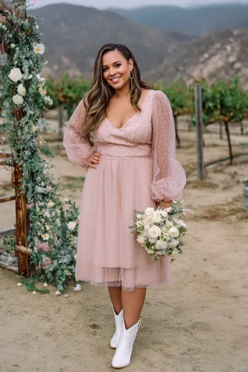 A curvy woman in a blush pink tulle midi dress with a lace bodice and sheer puff sleeves, paired with white cowboy boots
