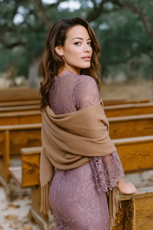 A curvy woman in a mauve lace midi dress with a scalloped hem and sheer sleeves, wrapped in a tan suede fringe shawl