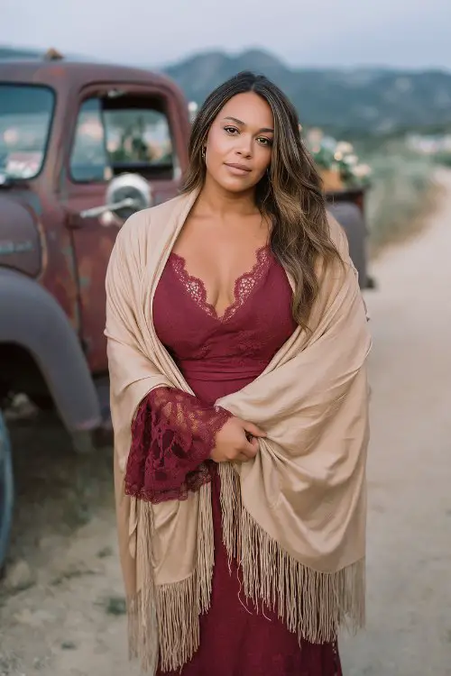 A curvy woman in a wine red boho dress with lace insets and bell sleeves, wrapped in a soft beige fringe shawl
