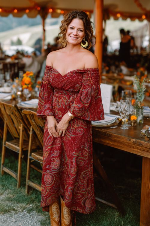 A curvy woman over 40 wears an oxblood red off-the-shoulder maxi dress with a bold paisley print, elegant flutter sleeves, and a soft wrap silhouette