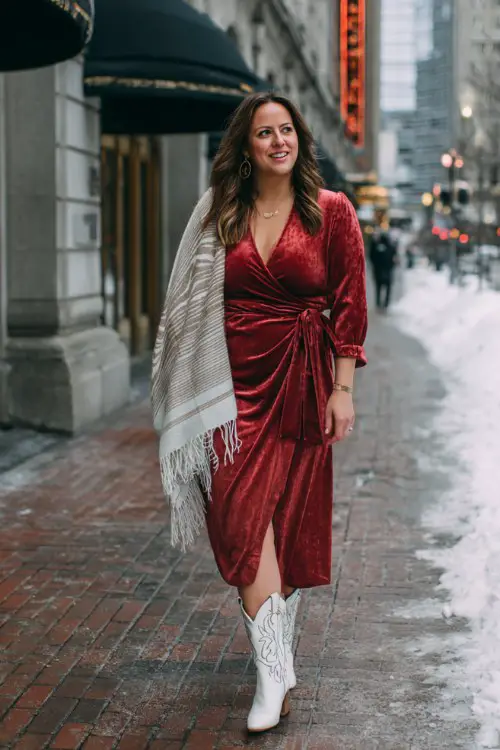 A curvy woman wears a deep red wrap midi dress with long sleeves, a matching velvet shawl, and white embroidered cowboy boots