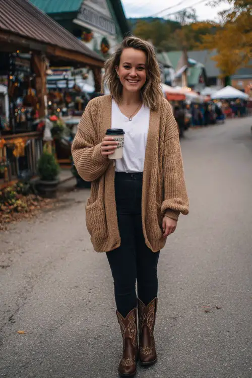 A curvy woman wears black skinny jeans, a camel-colored oversized cardigan, a tucked-in white tee, and embroidered cowboy boots