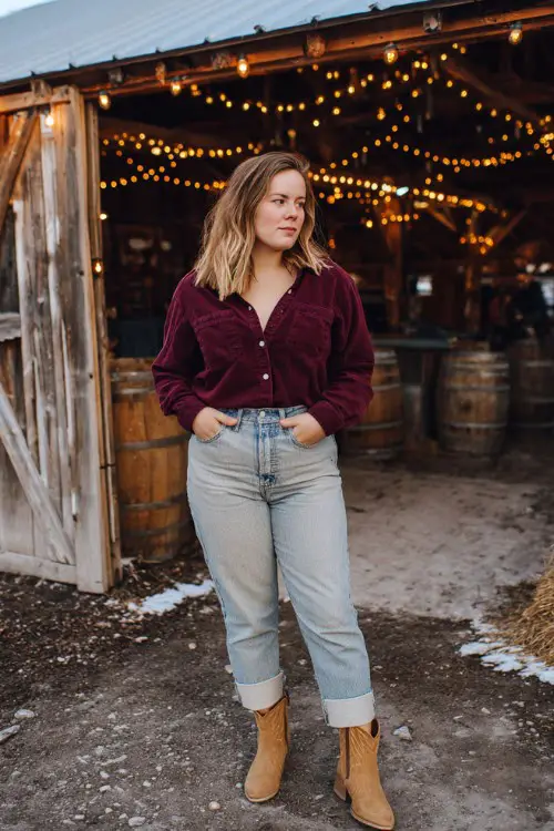 A curvy woman wears light-wash straight-leg jeans cuffed at the ankle, a burgundy button-up flannel, and tan short cowboy boots
