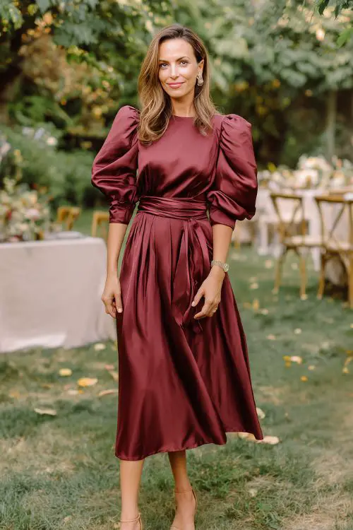 A fashionable woman over 40 in a tea-length burgundy satin dress with puff sleeves and pleated skirt