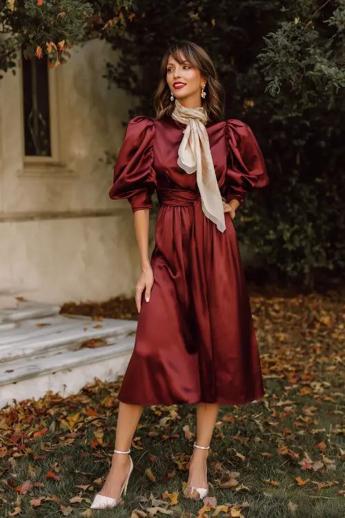 A fashionable woman over 50 in a tea-length burgundy satin dress with puff sleeves