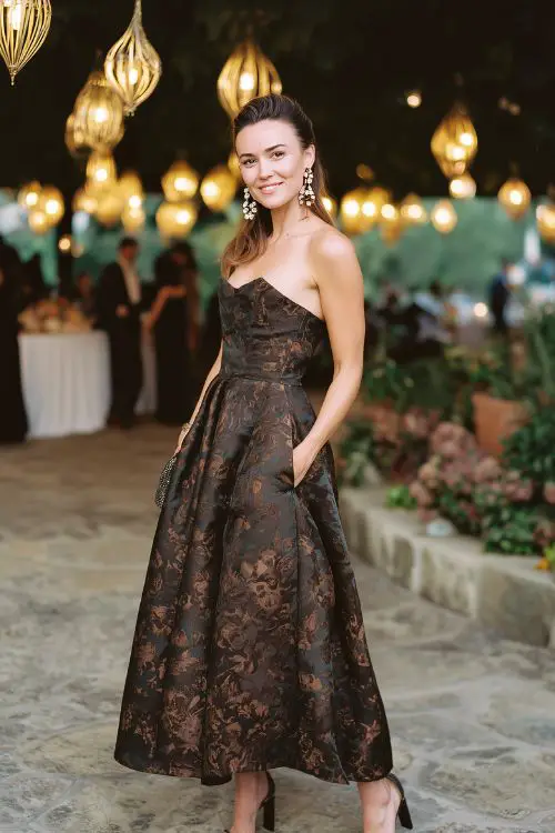 A guest in a dark floral jacquard gown with a fitted bodice and full-length skirt, styled with block heels and statement earrings