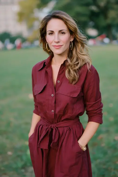 A guest in a maroon shirt dress with a tie waist and rolled-up sleeves, styled with flat leather boots and hoop earrings, attending a relaxed fall wedding picnic in the park