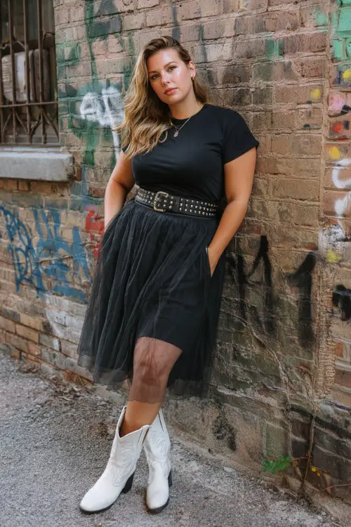 A plus-size woman wears a black tulle midi skirt with a fitted band tee tucked in, styled with white cowboy boots and a studded leather belt