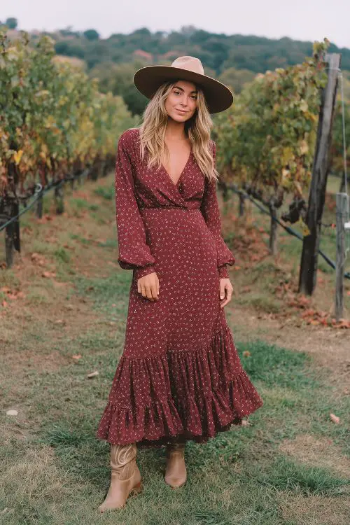 A stylish guest in a maroon maxi dress with small floral patterns, long sleeves, and a ruffle hem, styled with suede cowboy boots and a wide-brim hat