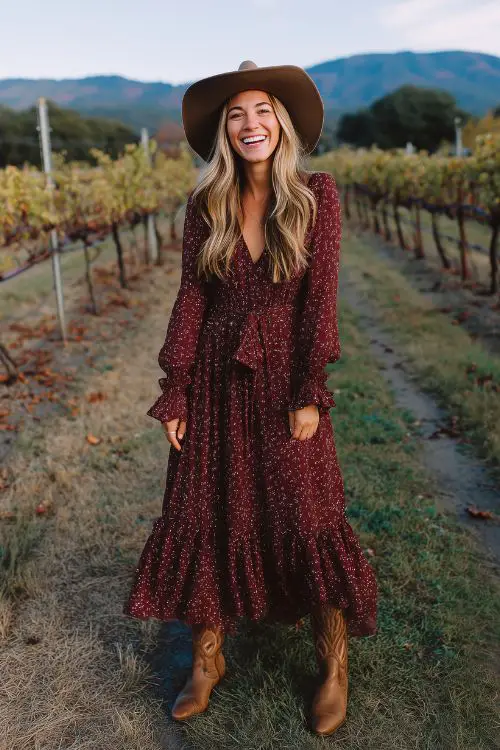 A stylish guest in a maroon maxi dress with small floral patterns, long sleeves, and a ruffle hem, styled with suede cowboy boots and a wide-brim hat