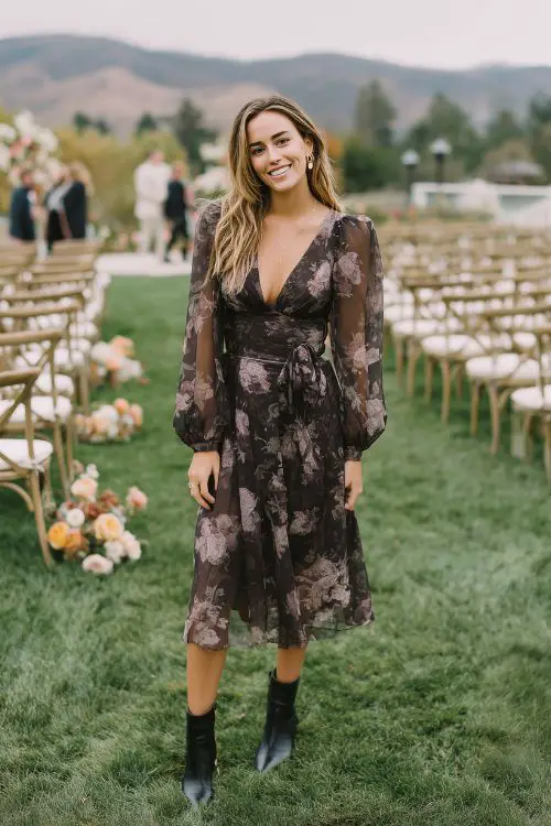 A wedding guest wears a dark floral midi dress with long chiffon sleeves and a cinched waist, paired with neutral-toned accessories and ankle boots