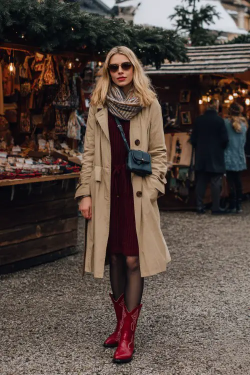 A woman browsing a winter market in a long camel trench coat over a maroon knit dress, black tights, and bold red cowboy boots