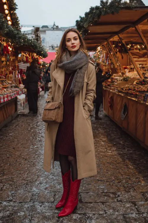 A woman browsing a winter market in a long camel trench coat over a maroon knit dress, black tights, and bold red cowboy boots