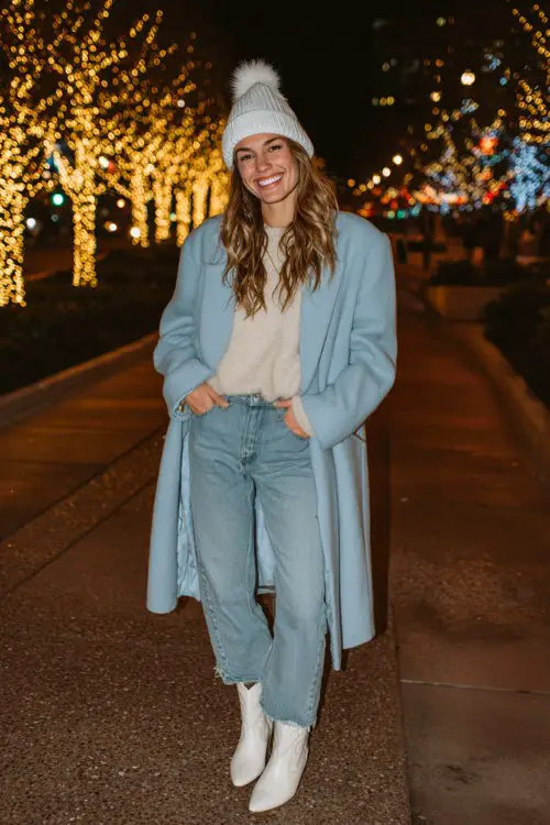 A woman enjoying holiday lights in a powder blue oversized coat, light wash jeans, thermal top, and pointed white cowboy boots