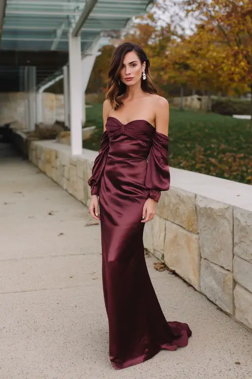A woman in a long burgundy satin gown with off-the-shoulder sleeves and a slight train, styled with pearl earrings and heeled pumps, standing near an outdoor venue surrounded by autumn leaves