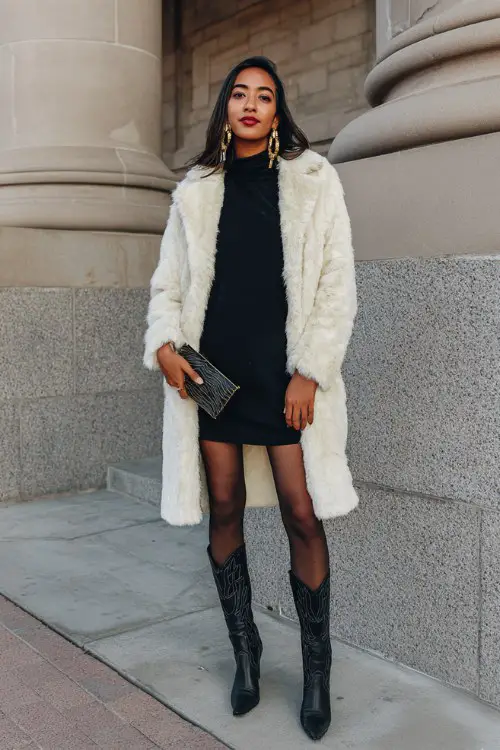 A woman in a long cream faux fur coat over a black turtleneck mini dress, sheer black tights, and tall black cowboy boots