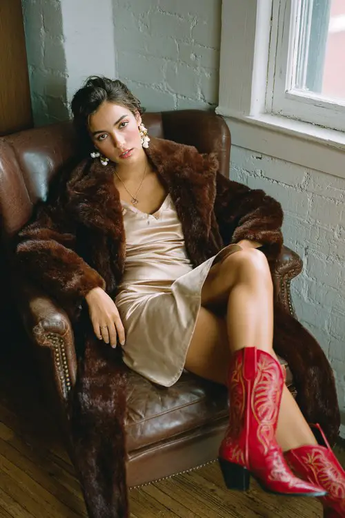 A woman lounging on a leather armchair in a chocolate faux fur coat, satin champagne slip dress, and red cowboy boots with embroidery, styled with pearl drop earrings