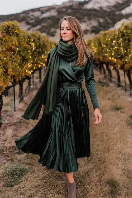 A woman over 30 in a forest green satin midi dress with a pleated skirt and scarf wrap