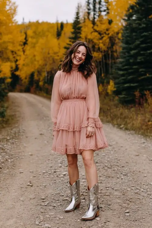 A woman over 30 wearing a blush pink A-line dress with a tiered sheer skirt and high neck, paired with metallic silver cowboy boots