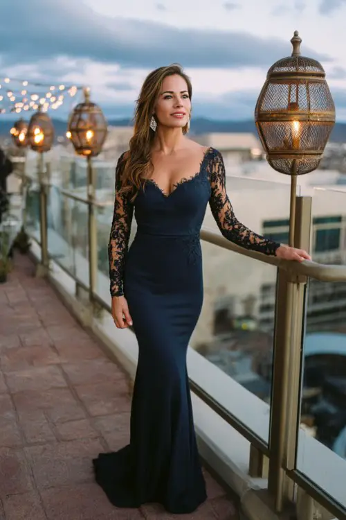 A woman over 30 wears a navy mermaid gown with sheer lace panels along the sleeves and sides
