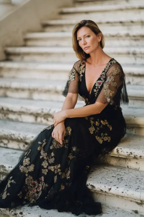 A woman over 40 in a floor-length black gown with metallic floral embroidery and sheer cape sleeves