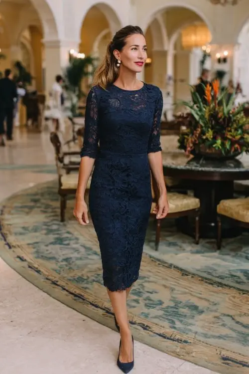 A woman over 40 wears a navy lace sheath dress with three-quarter sleeves and pearl earrings