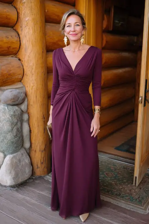 A woman over 50 wearing a plum-colored jersey maxi dress with a defined waist and elegant drape, paired with pointed-toe flats and gold drop earrings