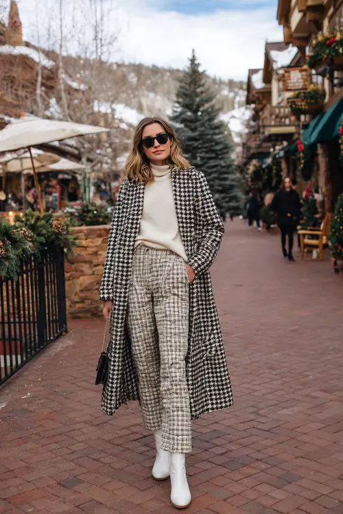 A woman shopping in a quaint winter village wearing a houndstooth coat, wide-leg wool trousers, a cashmere sweater, and structured white cowboy boots