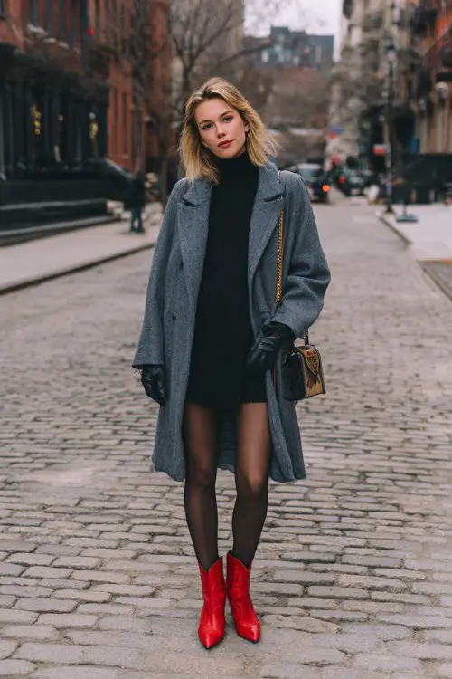 A woman standing on a cobblestone street in a gray oversized wool coat, black turtleneck dress, sheer tights, and striking red cowboy boots
