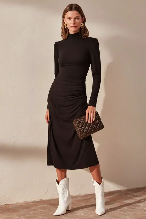 A woman wears a black long-sleeve midi dress with a high neckline, elegant darting, and a soft flare, paired with structured white cowboy boots and a quilted leather clutch