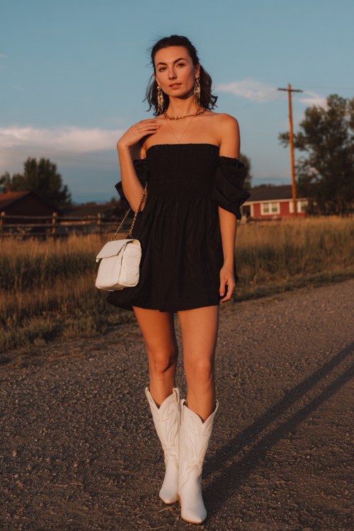 A woman wears a black off-shoulder dress with a flowy silhouette and white cowboy boots, styled with statement earrings