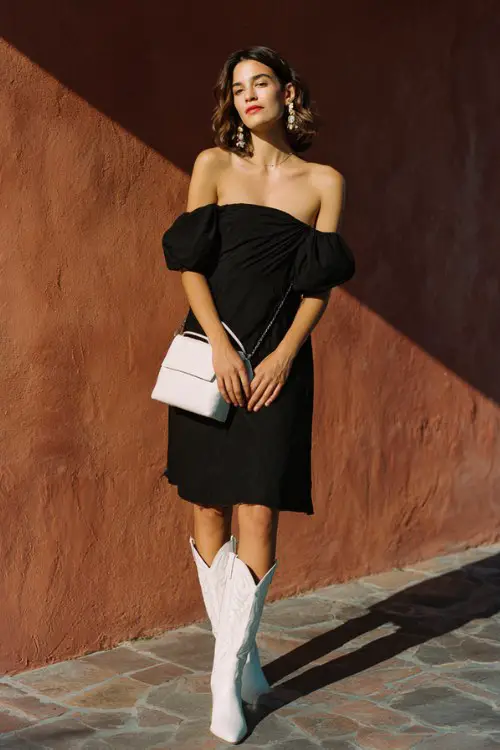 A woman wears a black off-shoulder dress with a flowy silhouette and white cowboy boots