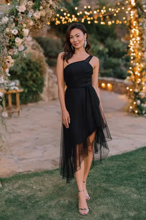 A woman wears a black one-shoulder midi dress with an asymmetrical layered hem and sheer overlay (2)