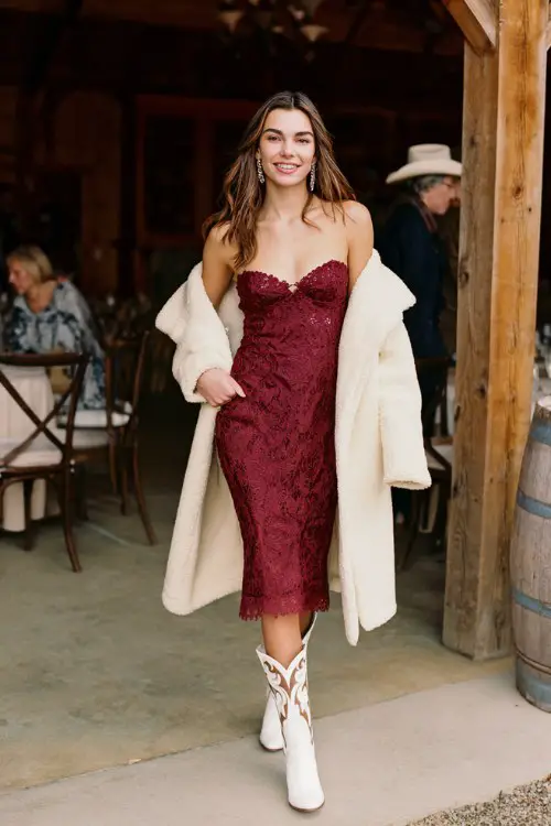 A woman wears a burgundy lace-trimmed midi dress with a sweetheart neckline, a cream wool coat draped over her shoulders, and white cowboy boots